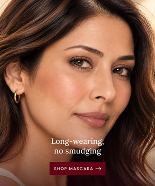 Close-up beauty ad showing a woman with long brown hair and natural makeup, wearing a small gold hoop earring. White text reads 'Long-wearing, no smudging' with a dark red button below that says 'SHOP MASCARA →'.